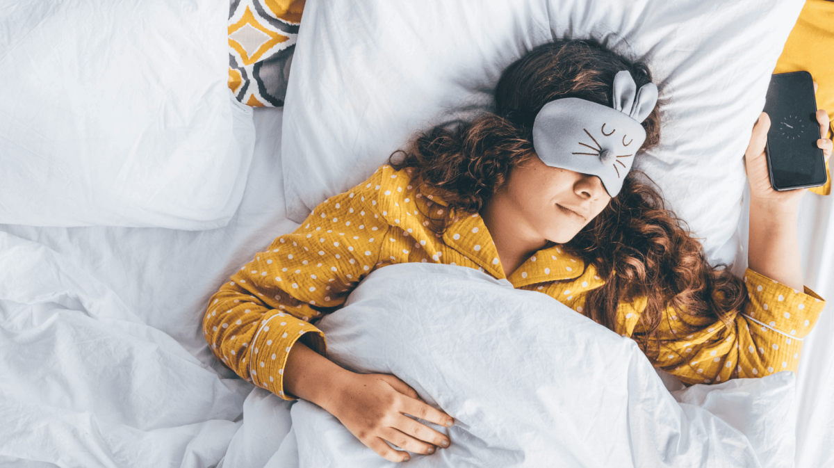 Millennial-Frau im Bett liegend mit Schlafmaske und Telefon in der Hand.