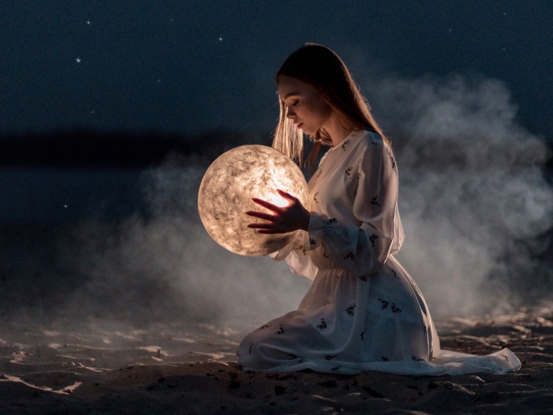 Frau sitzt an Strand und hält Mond in der Hand.