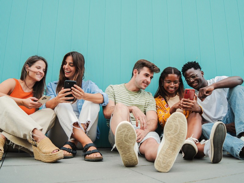 Fünf Freunde auf dem Boden sitzend vor einer türkisen Wand, die sich unterhalten und etwas auf dem Handy Display zeigen