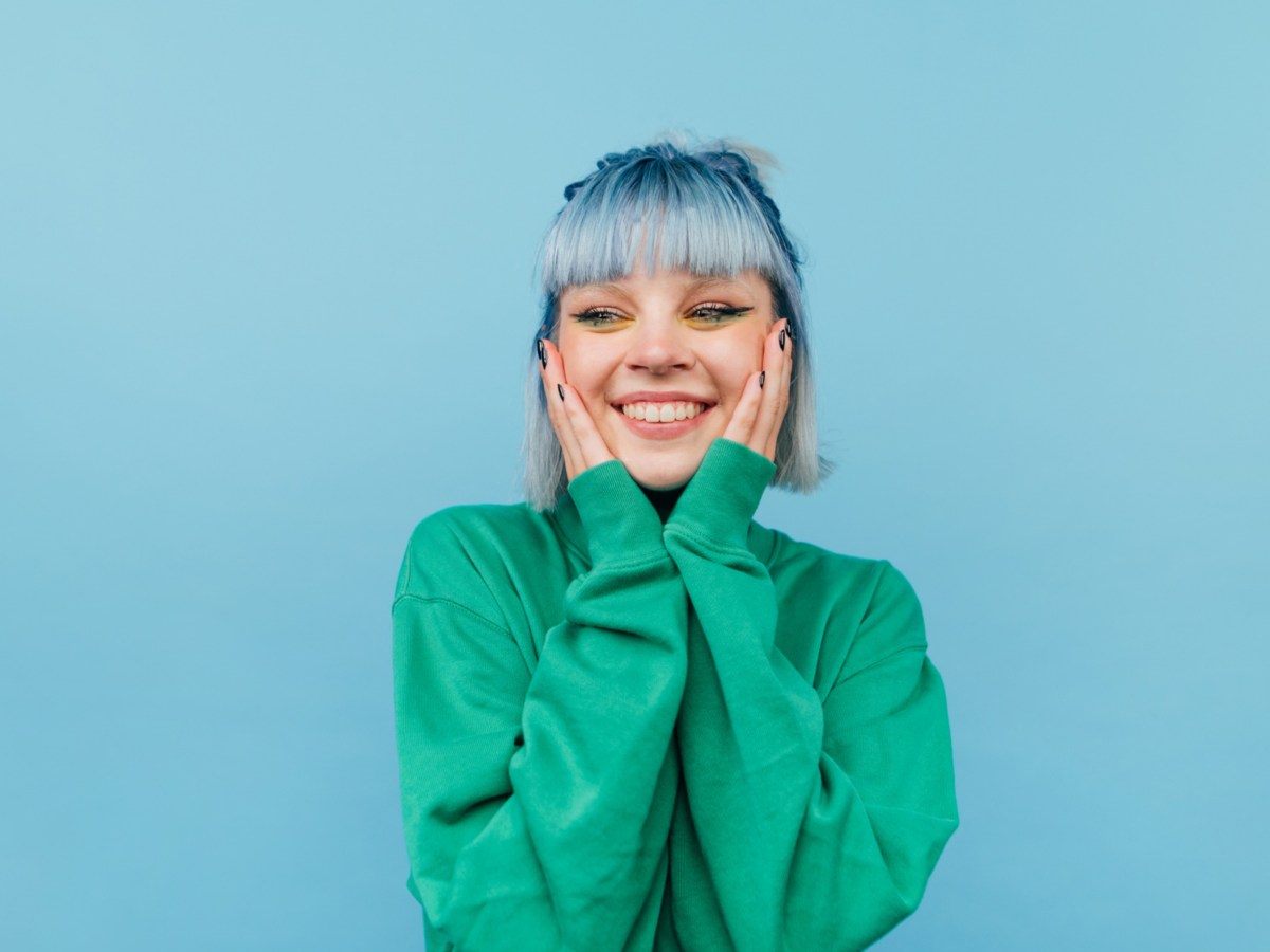 Frau mit blauen Haaren vor blauer Wand, die sich mit den Händen an die Wange fasst und lächelt