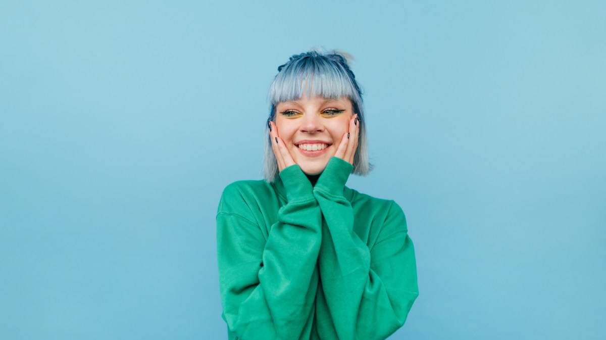 Frau mit blauen Haaren vor blauer Wand, die sich mit den Händen an die Wange fasst und lächelt