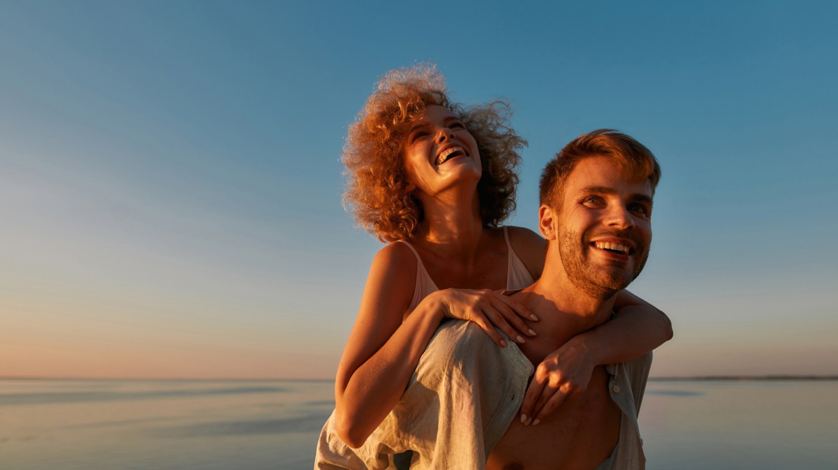 Frau, die Huckepack auf einem Mann am Strand ist, mit dem Sonnenuntergang im Hintergrund, während beide lächeln