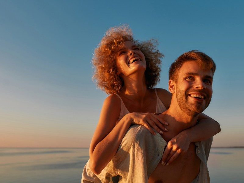 Frau, die Huckepack auf einem Mann am Strand ist, mit dem Sonnenuntergang im Hintergrund, während beide lächeln