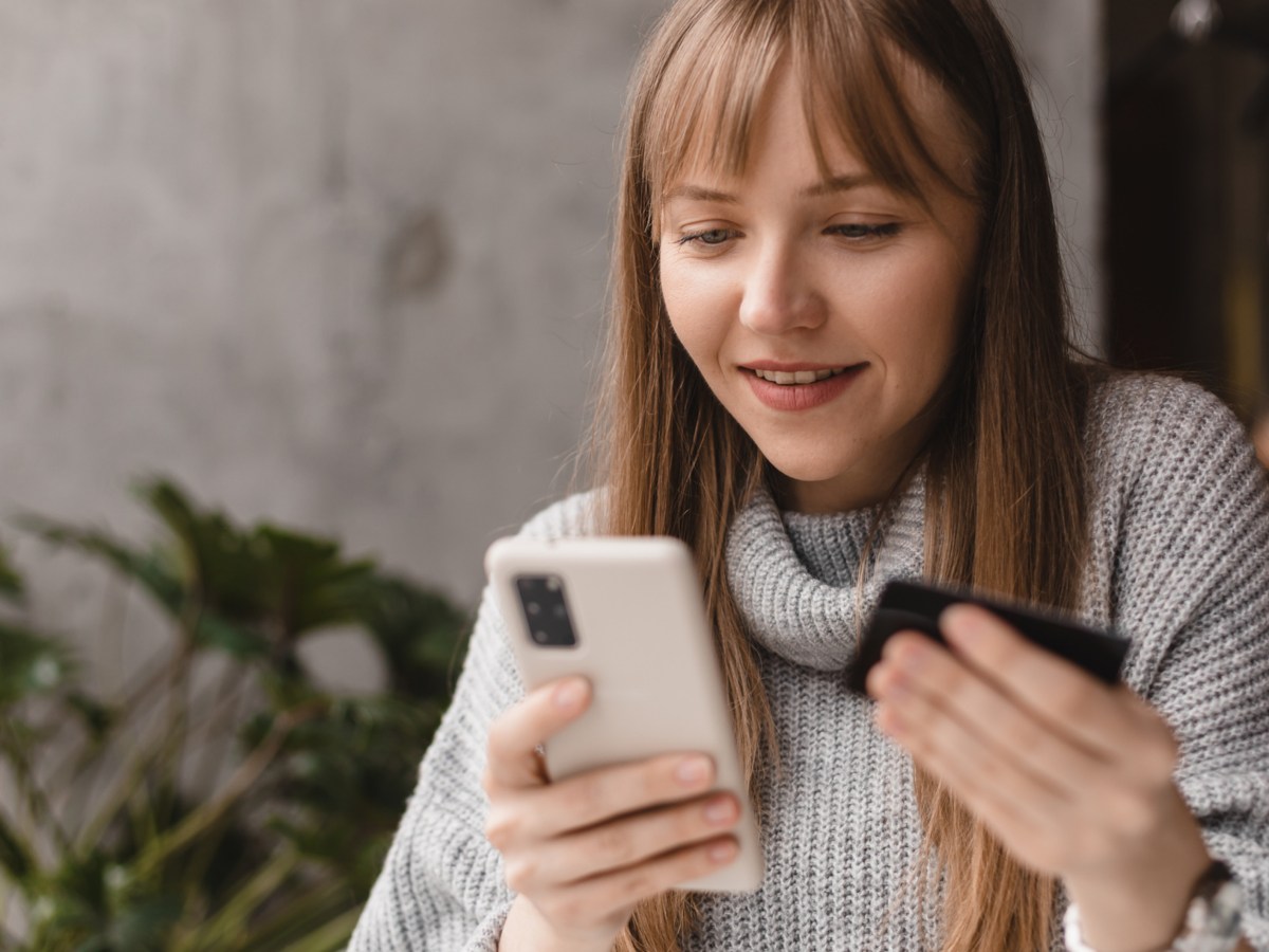 Junge Frau hält eine Kreditkarte in der Hand und ihr Handy in der anderen Hand und schaut gespannt auf das Telefon