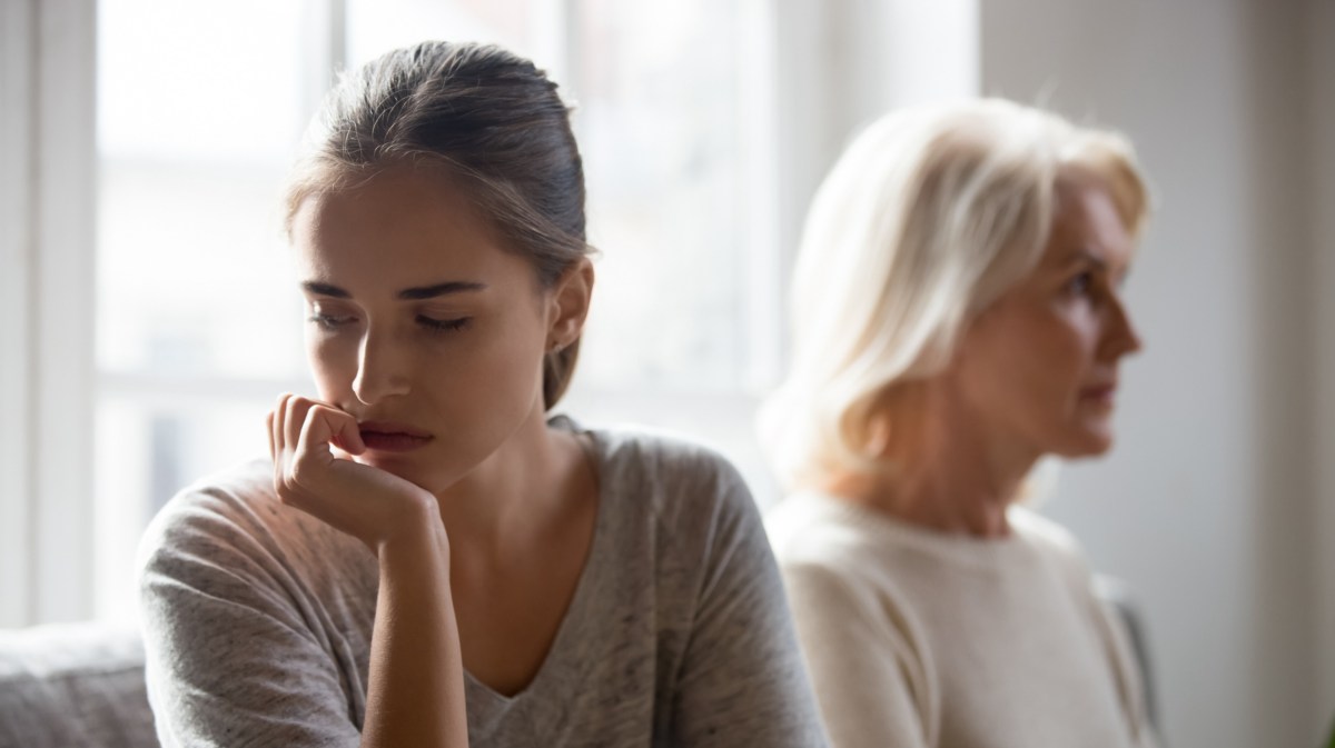 Junge Frau, die auf dem Sofa sitzt , mit Hand an ihrem Kinn abgestützt, während eine ältere Frau hinter ihr verschwommen sitzt