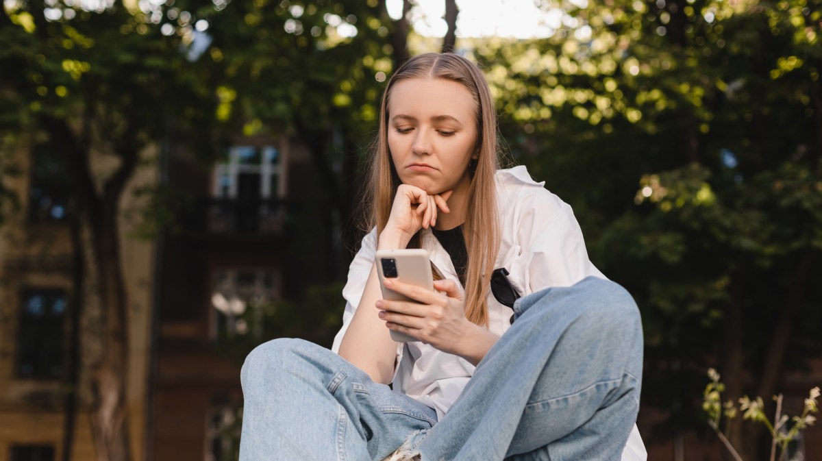 Frau in Jeans, die in einem Park sitzt und ihr Handy fragwürdig anschaut