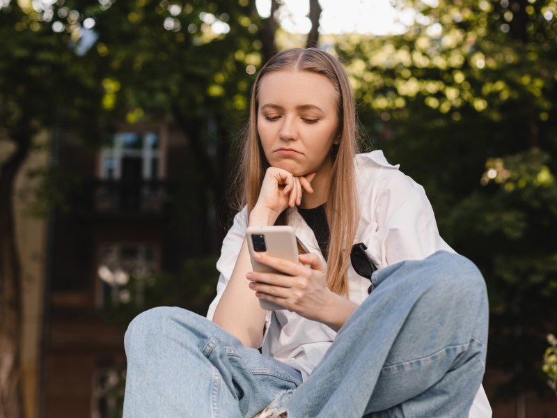 Frau in Jeans, die in einem Park sitzt und ihr Handy fragwürdig anschaut