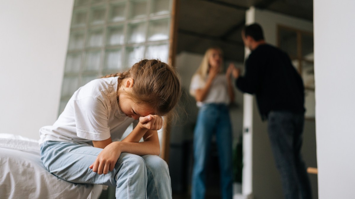 Zwei Erwachsene, die sich im Hintergrund streiten, während ein Kind im Vordergrund ihre Hände vors Gesicht nimmt