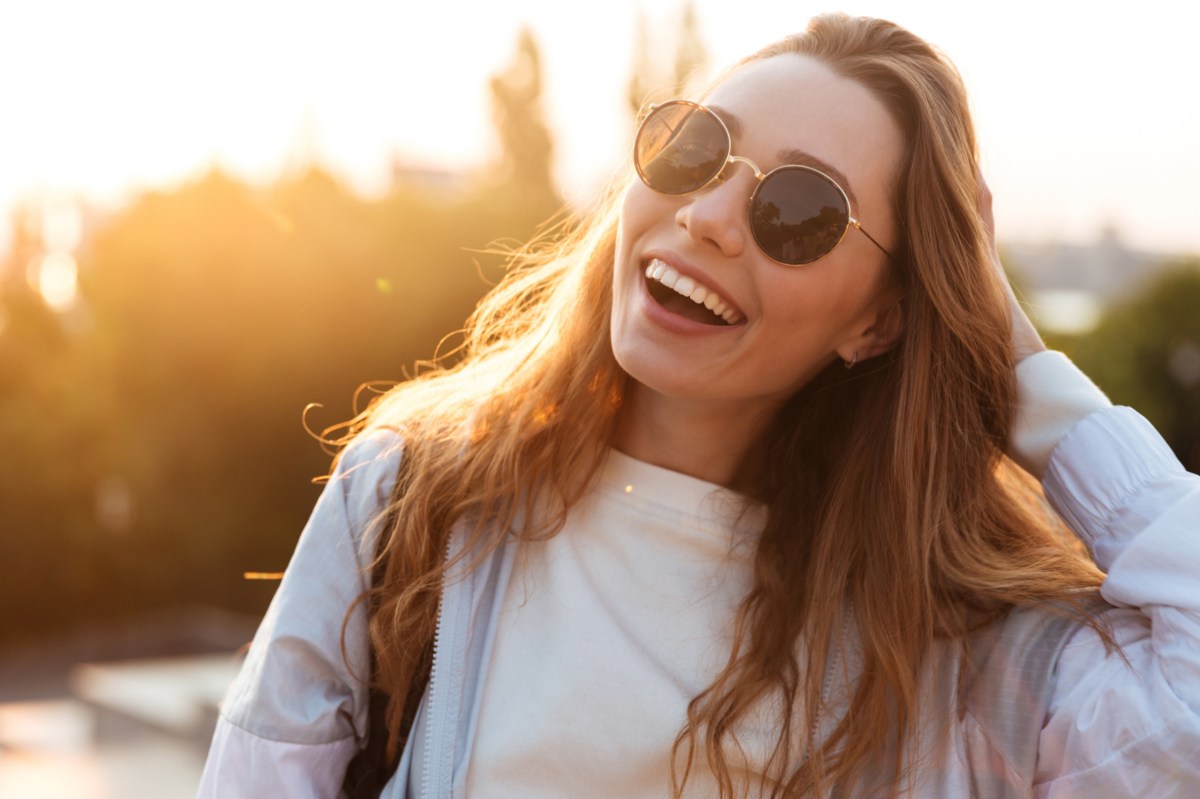 Frau mit Sonnenbrille geht sich durch die Haare und steht lachend in der Sonne.