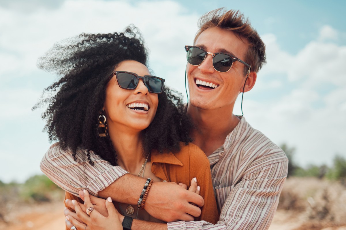 Mann mit Sonnenbrille umarmt Frau mit Sonnenbrille. Beide sind ein Paar und lachen gemeinsam.