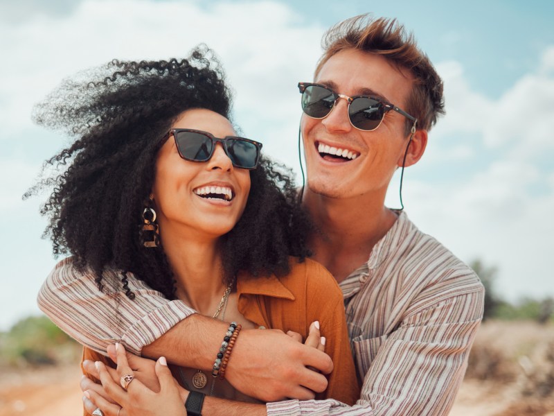 Mann mit Sonnenbrille umarmt Frau mit Sonnenbrille. Beide sind ein Paar und lachen gemeinsam.