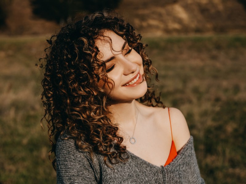 Frau mit lockigem Haar steht in der Sonne auf einer Wiese und lächelt.