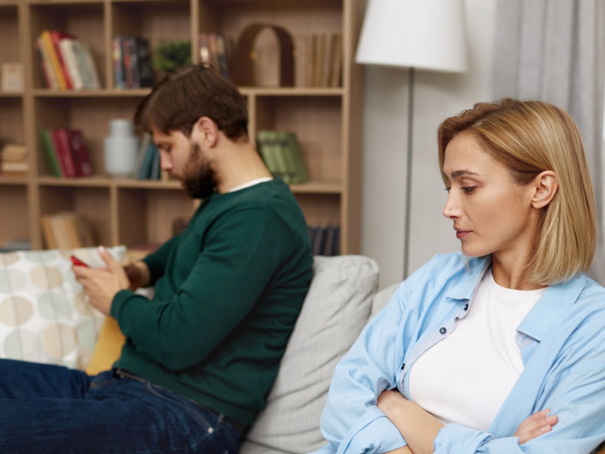 Frau mit blonden Haaren, die mit einegschränkten Armen auf der Couch sitzt und in Richtung ihres Partners schaut, der im Hintergrund abweisend auf den Boden guckt