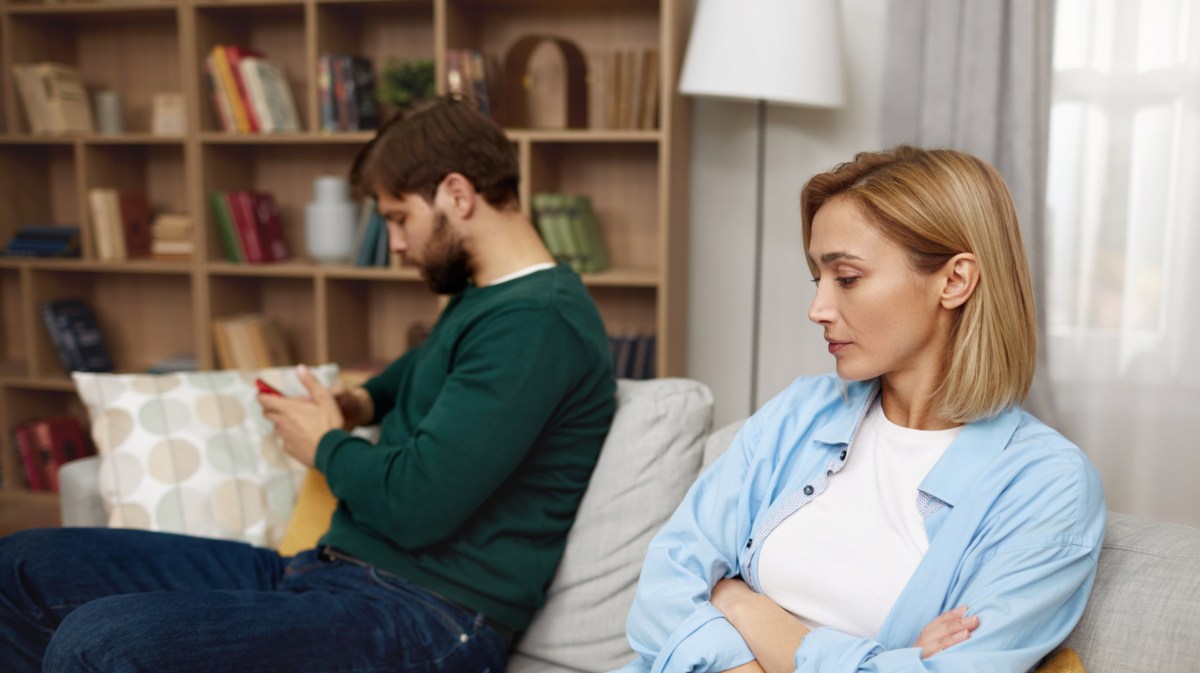 Frau mit blonden Haaren, die mit einegschränkten Armen auf der Couch sitzt und in Richtung ihres Partners schaut, der im Hintergrund abweisend auf den Boden guckt