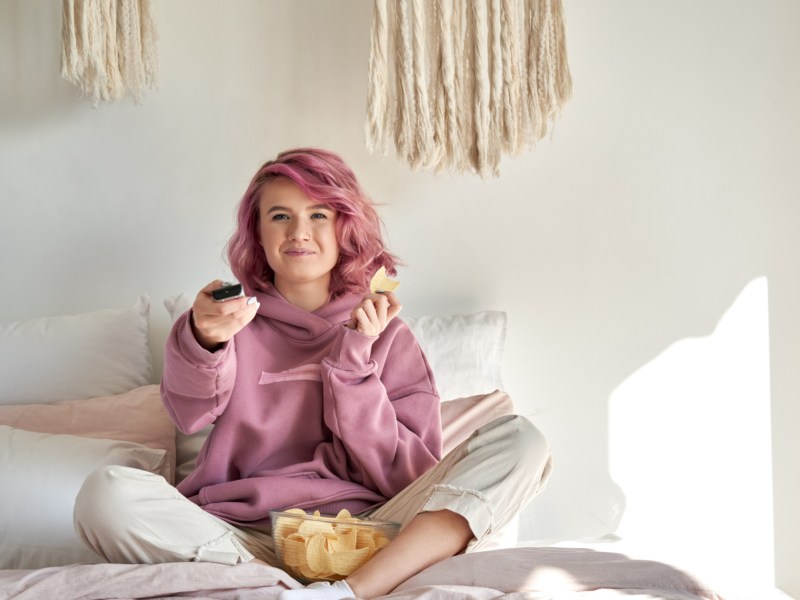 Frau mit rosa Haaren auf ihren Bett, die Chips isst und eine Fernbedienung in der Hand hält