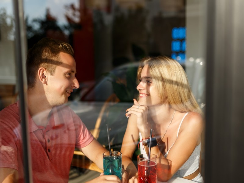 Mann und Frau auf einem Date mit zwei Drinks, hinter einer Glasscheibe, die sich gegenseitig anlächeln