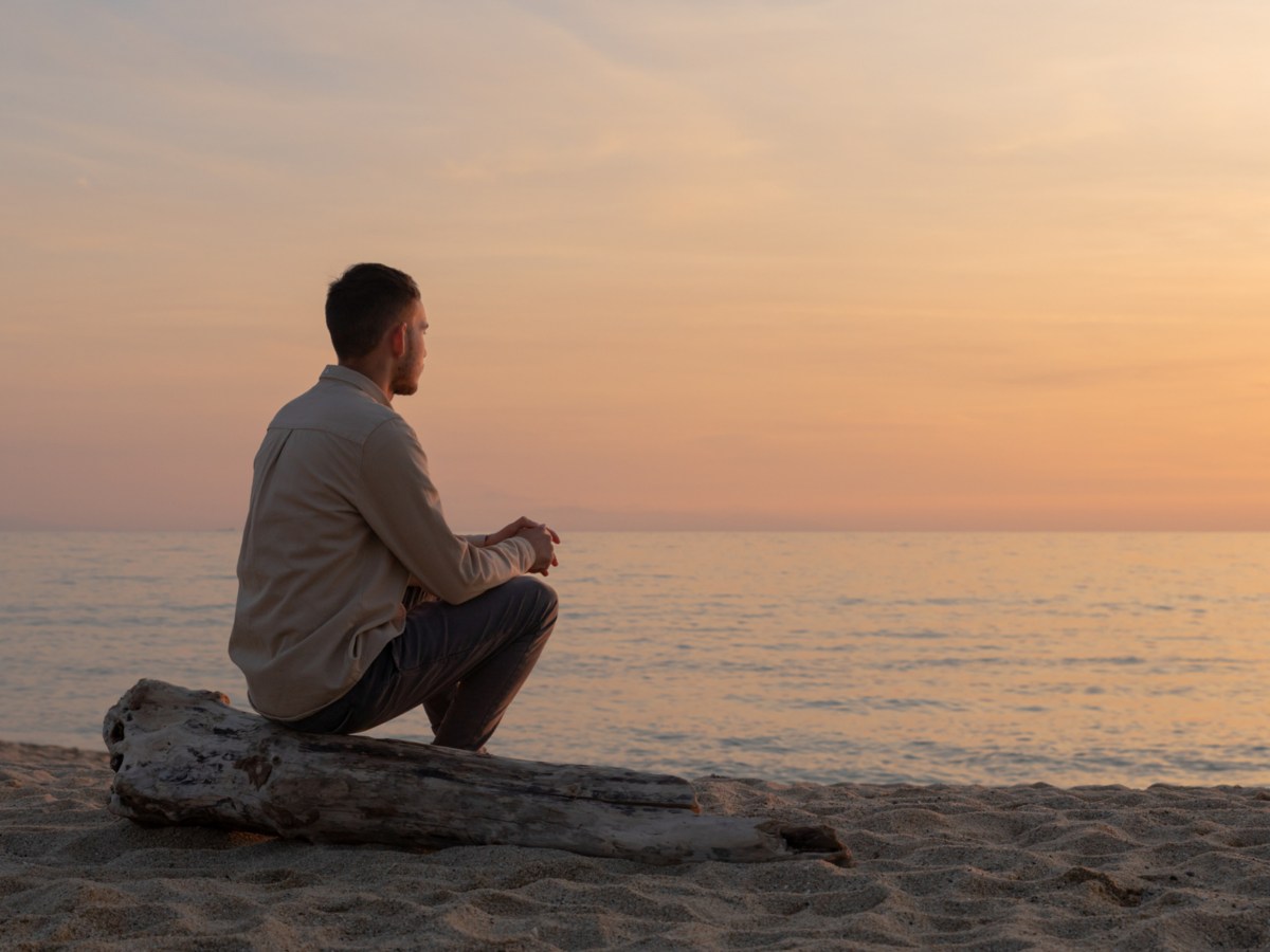 Mann am Strand, der mit dem R&uuml;cken zur Kamera sitzt und auf das Meer schaut im Sonnenuntergang