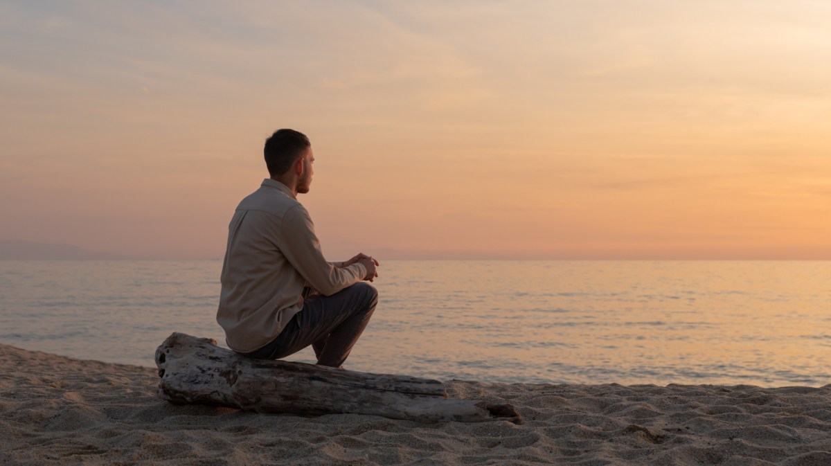 Mann am Strand, der mit dem Rücken zur Kamera sitzt und auf das Meer schaut im Sonnenuntergang