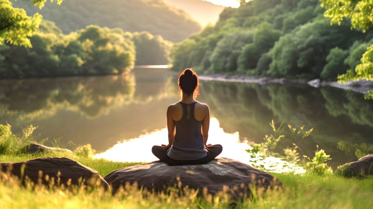 Eine Frau sitzt auf einem Stein am See und meditiert.