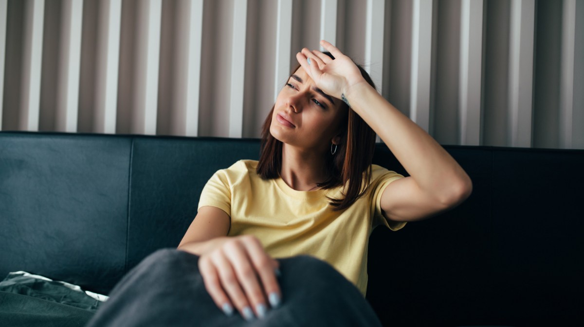 Frau in gelbem Shirt, die auf der Couch sitzt und sich die Handfläche auf die Stirn legt und dabei die Stirn runzelt