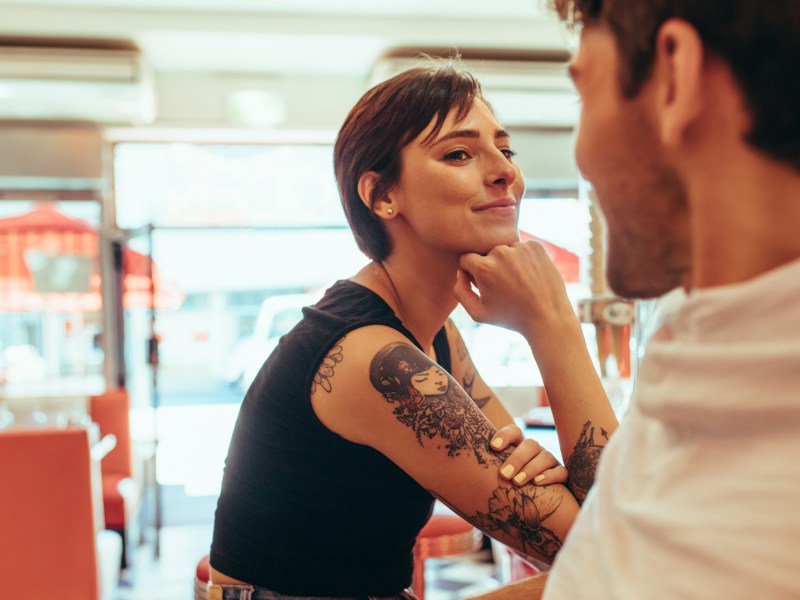 Frau mit Tattoos und kurzen Haaren in einem Waschsalon, die einen Mann gegenüber anlächelt und flirtet