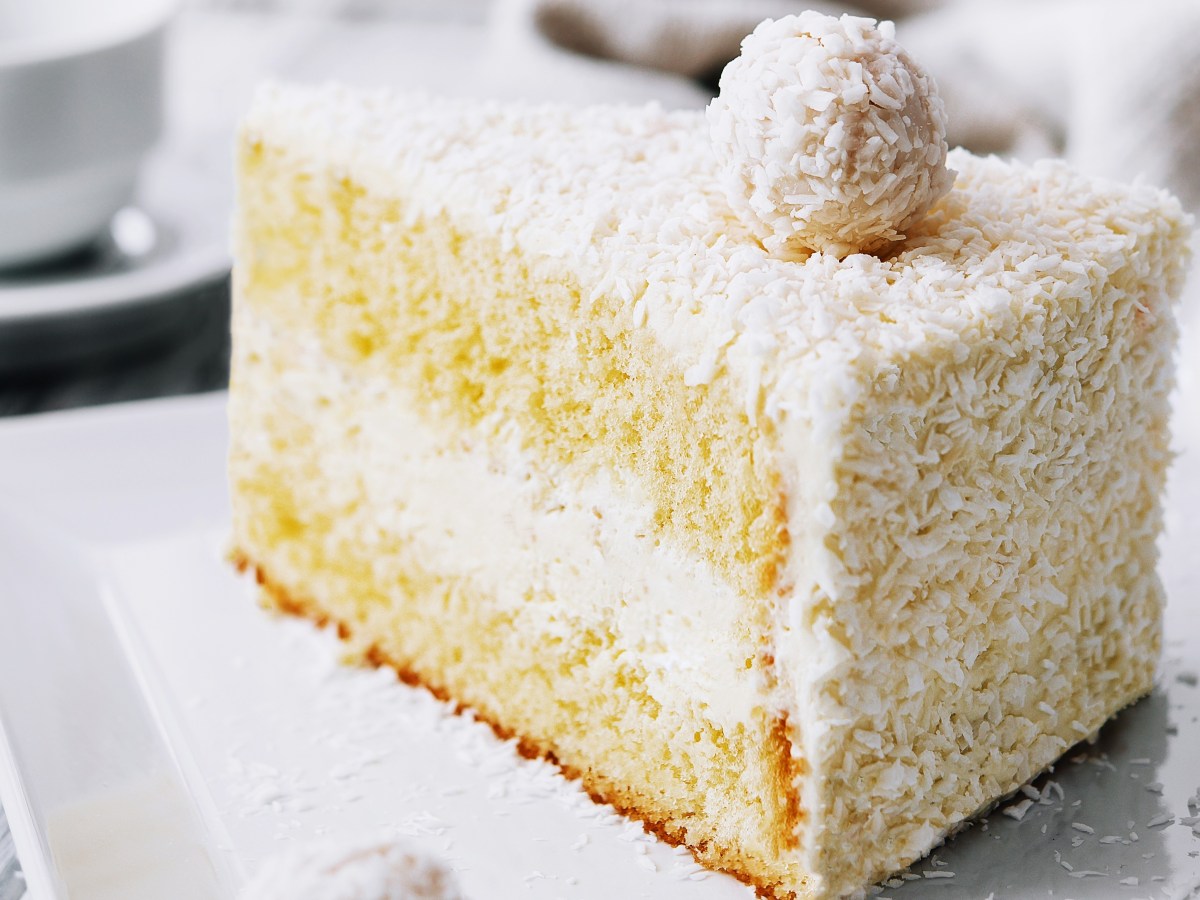 Ein Stück vom weißen Raffaello-Kuchen auf einem weißen Teller und hellem Holztisch.