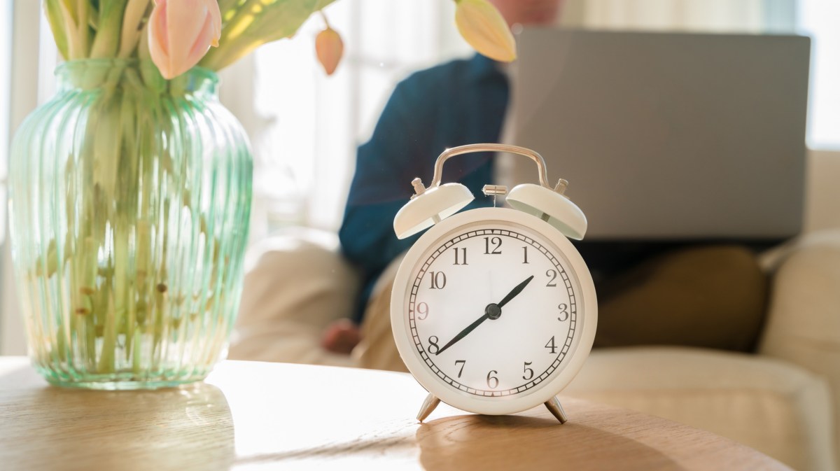 Mann mit Laptop auf dem Schoß, der im Hintergrund sitzt, wo im Vordergrund eine Uhr mit einer Vase auf dem Tisch steht