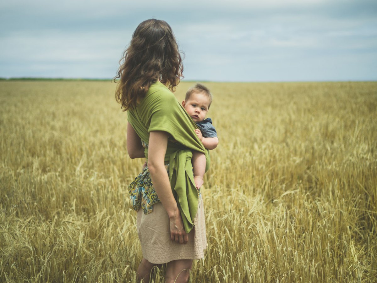 Eine junge Frau tr&auml;gt ihr Kind in einem Tuch und steht auf einer gr&uuml;nen Wiese