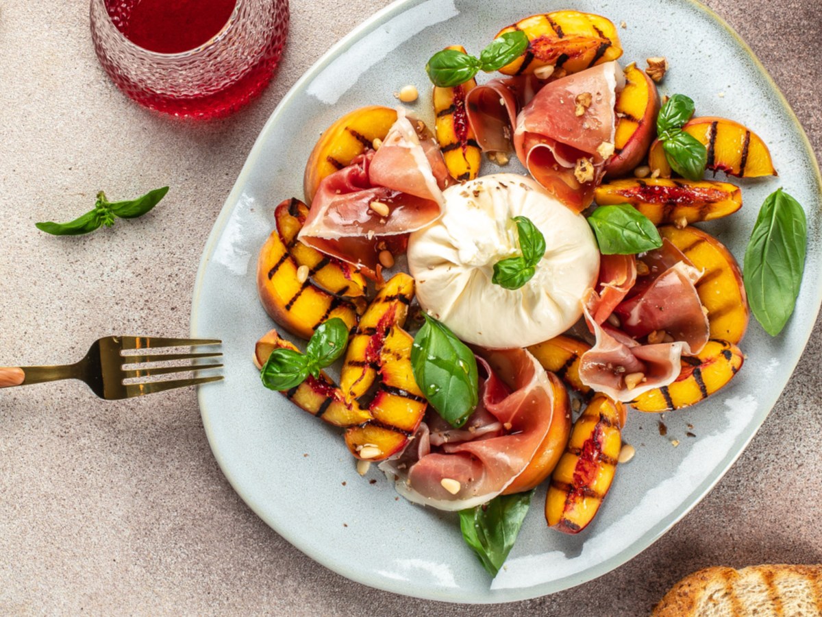 Pfirsich, Burrata, Schinken und Basilikum auf einem Teller. Daneben stehen Getränke und geröstetes Brot.