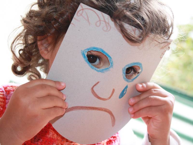 Kind hält sich eine traurige, selbstgemalte Maske vors Gesicht.