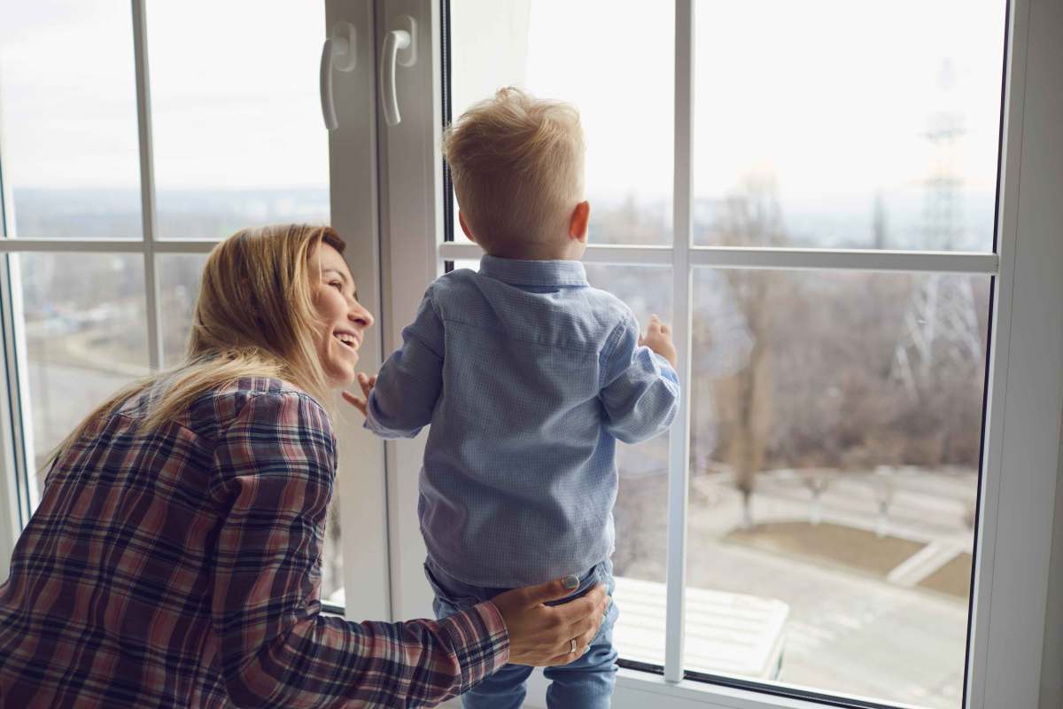 Mutter hält ihren ca. 2-jährigen Sohn fest, während der auf dem Fensterbrett steht und nach draußen schaut.