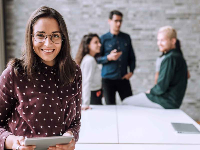 Junge Frau mit braunen Haaren und Brille lächelt in gemusterter Bluse mit Tablet, im Hintergrund drei Kollegen in lockerer Unterhaltung.