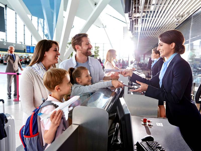 Vierköpfige Familie steht am Check-in-Schalter eines Flughafens und reicht der Mitarbeiterin die Reisepässe.