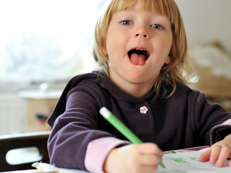 Kleinkind beim Malen an einem Schreibtisch, schaut in die Kamera und streckt die Zunge raus..