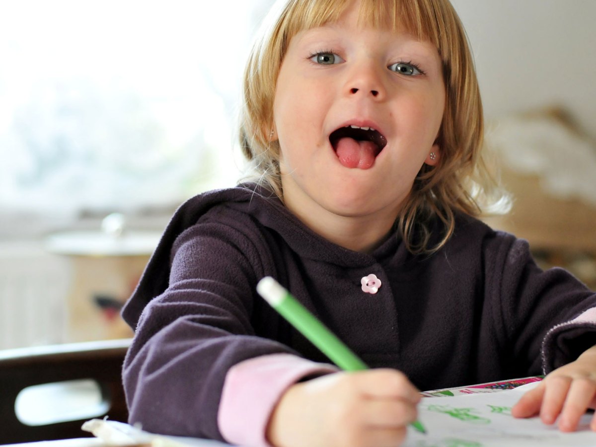 Kleinkind beim Malen an einem Schreibtisch, schaut in die Kamera und streckt die Zunge raus..