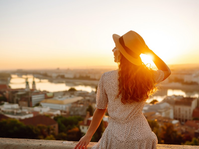 Frau mit Hut sitzt auf einer Mauer vor einer traumhaften Stadtkulisse im Süden bei einem Sonnenuntergang.