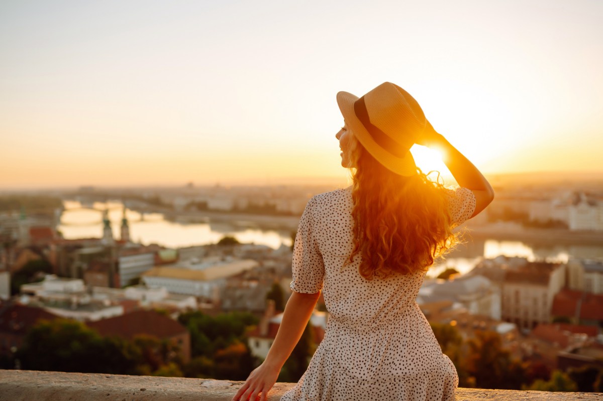 Frau mit Hut sitzt auf einer Mauer vor einer traumhaften Stadtkulisse im Süden bei einem Sonnenuntergang.