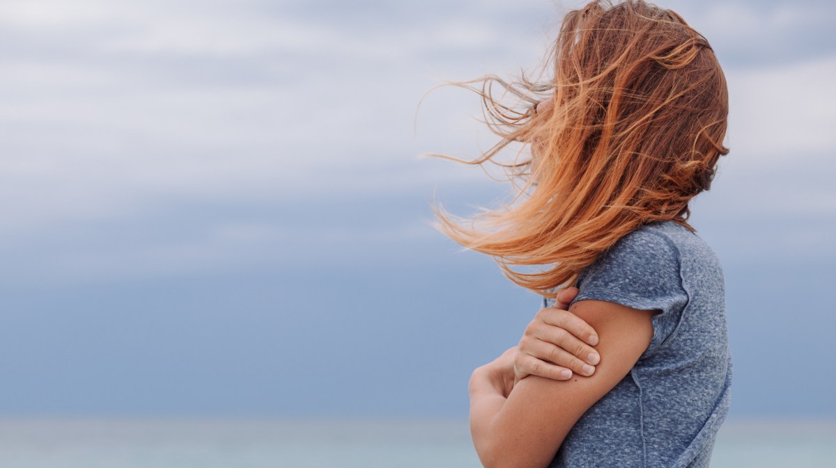 Frau mit blauem Shirt vor blauem Hintergrund umarmt sich selbst.