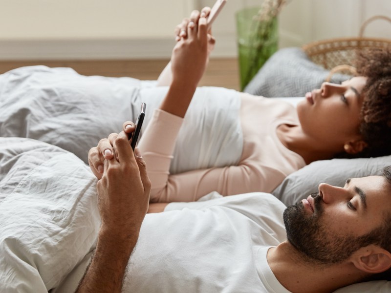 Mann und Frau die gemeinsam nebeneinander im Bett liegen und gelangweilt auf ihr Handy schauen