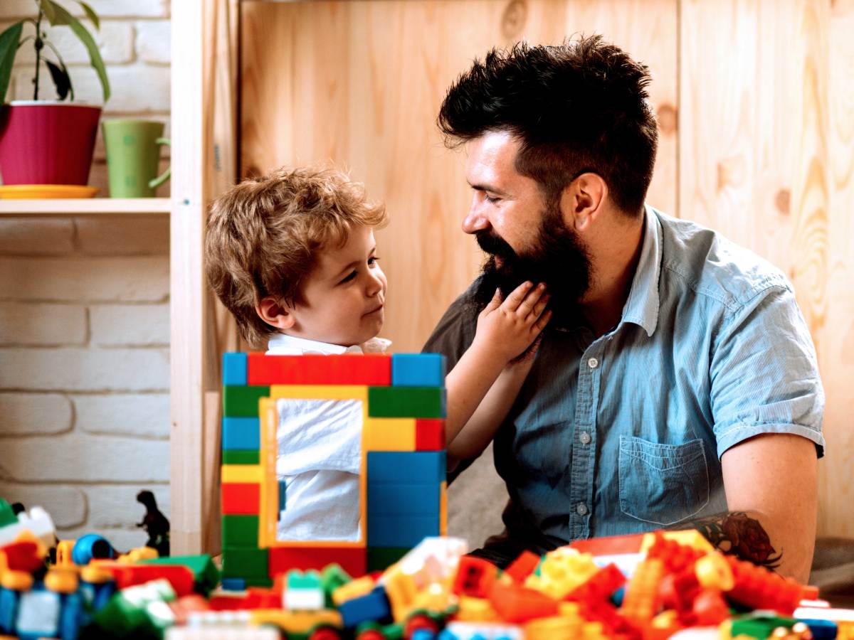 Vater und Sohn sitzen vor einem Stapel Bausteinen. Der Sohn fasst gerade in den Vollbart seines Vaters.