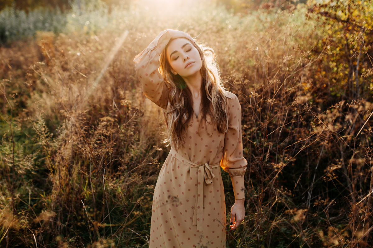 Frau in einem Feld voller Gras, die ein Kleid trägt und ihren Kopf zur Seite zieht, während die Sonne von hinten auf sie scheint