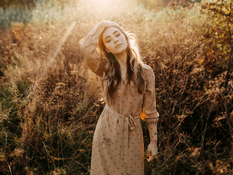 Frau in einem Feld voller Gras, die ein Kleid trägt und ihren Kopf zur Seite zieht, während die Sonne von hinten auf sie scheint