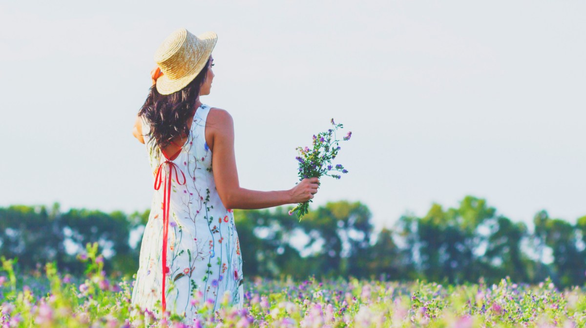 Eine Frau steht mit Kleid und Hut in einem Blumenfeld.