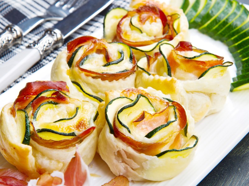 Blätterteig-Rosen gefüllt mit Zucchini, Käse und Schinken auf einem weißen Teller.