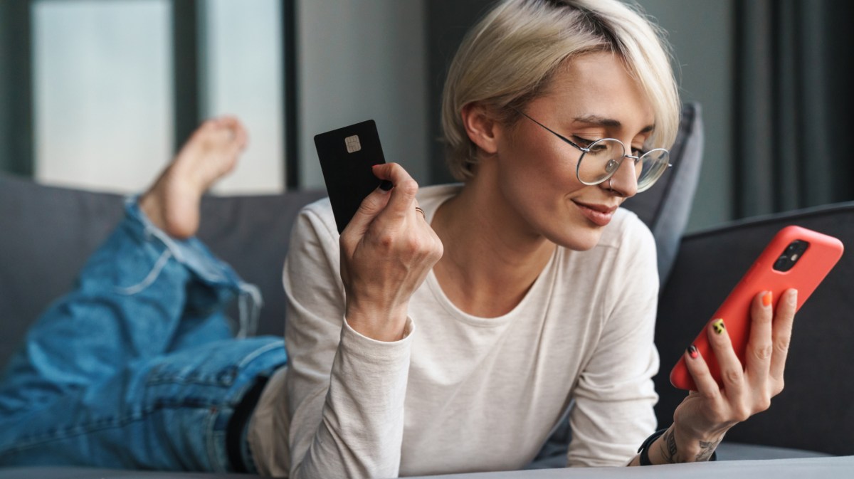 Frau mit kurzen Haaren liegt auf dem Sofa und hat Kreditkarte und Handy in der Hand.