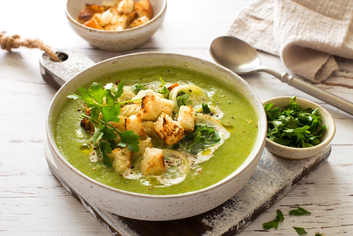 Eine Schüssel Zucchini-Spinat-Suppe mit Croûtons und frischer Petersilie garniert, daneben eine Schüssel mit gehackter Petersilie und eine mit Croûtons, außerdem ein Löffel und ein Geschirrtuch.