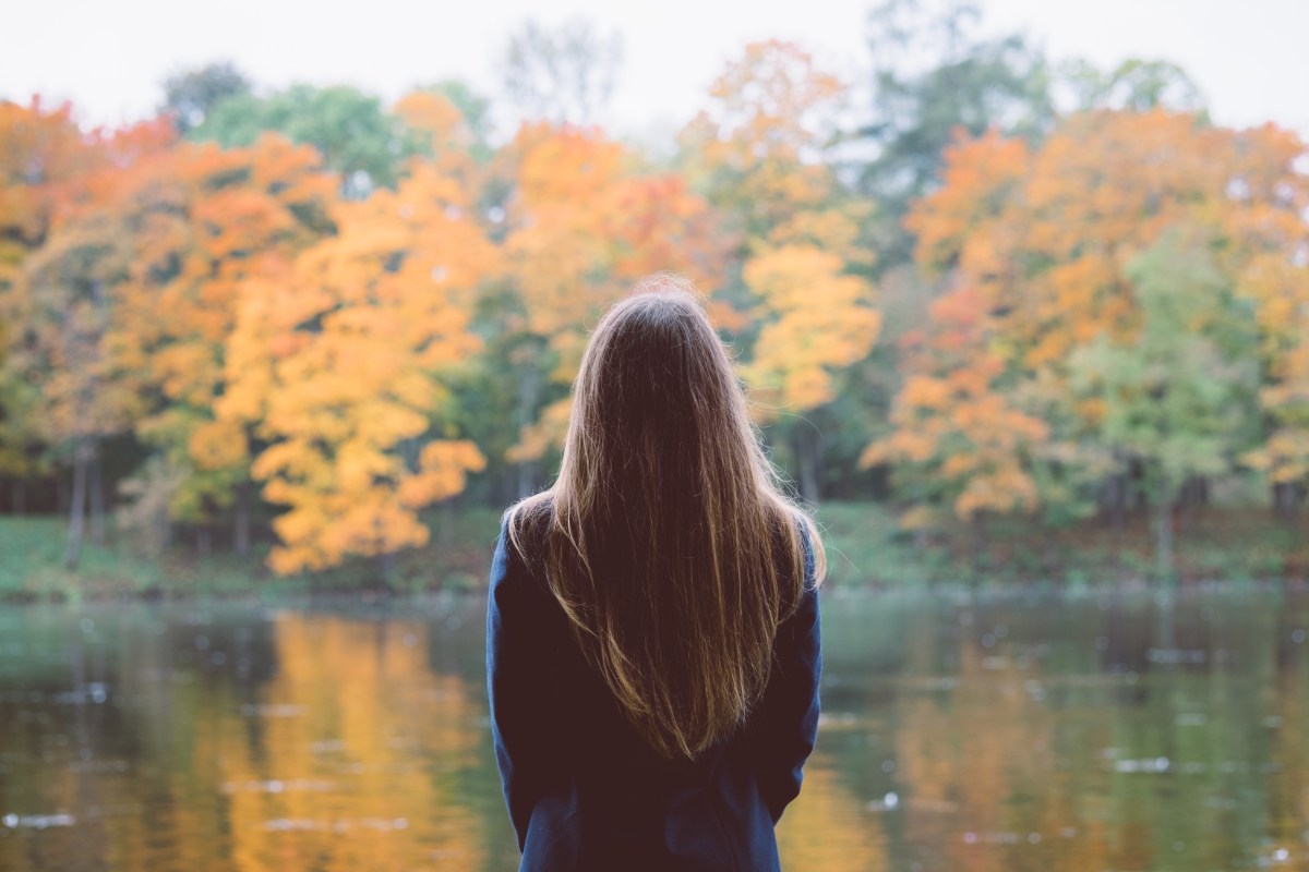 Junge Frau mit braunen Haaren steht vor einem See im Herbst und hat der Kamera den Rücken zugwandt.