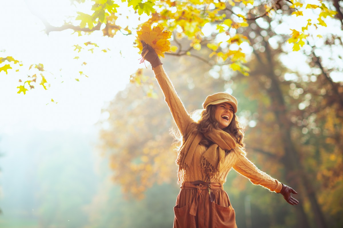 Frau im Herbstoutfit, mit gelben Blättern in der Hand im herbstlichen Wald