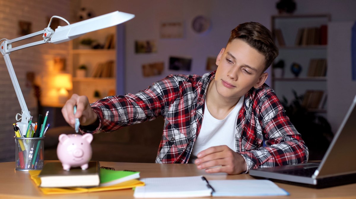 Teenager sitzt an seinem Schreibtisch und schiebt einen Geldschein in sein Sparschwein.