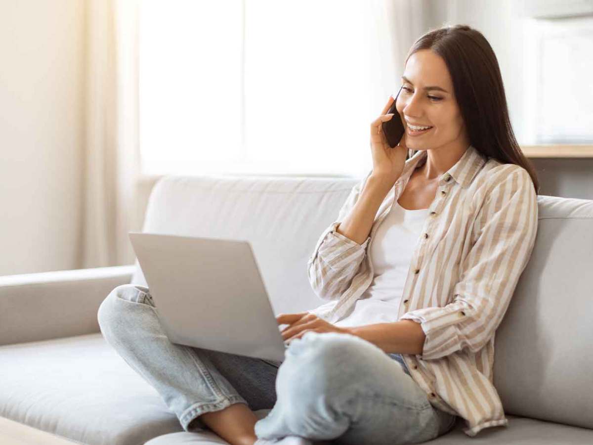 Frau sitzt zu Hause mit Laptop und Handy auf der Couch und arbeitet.